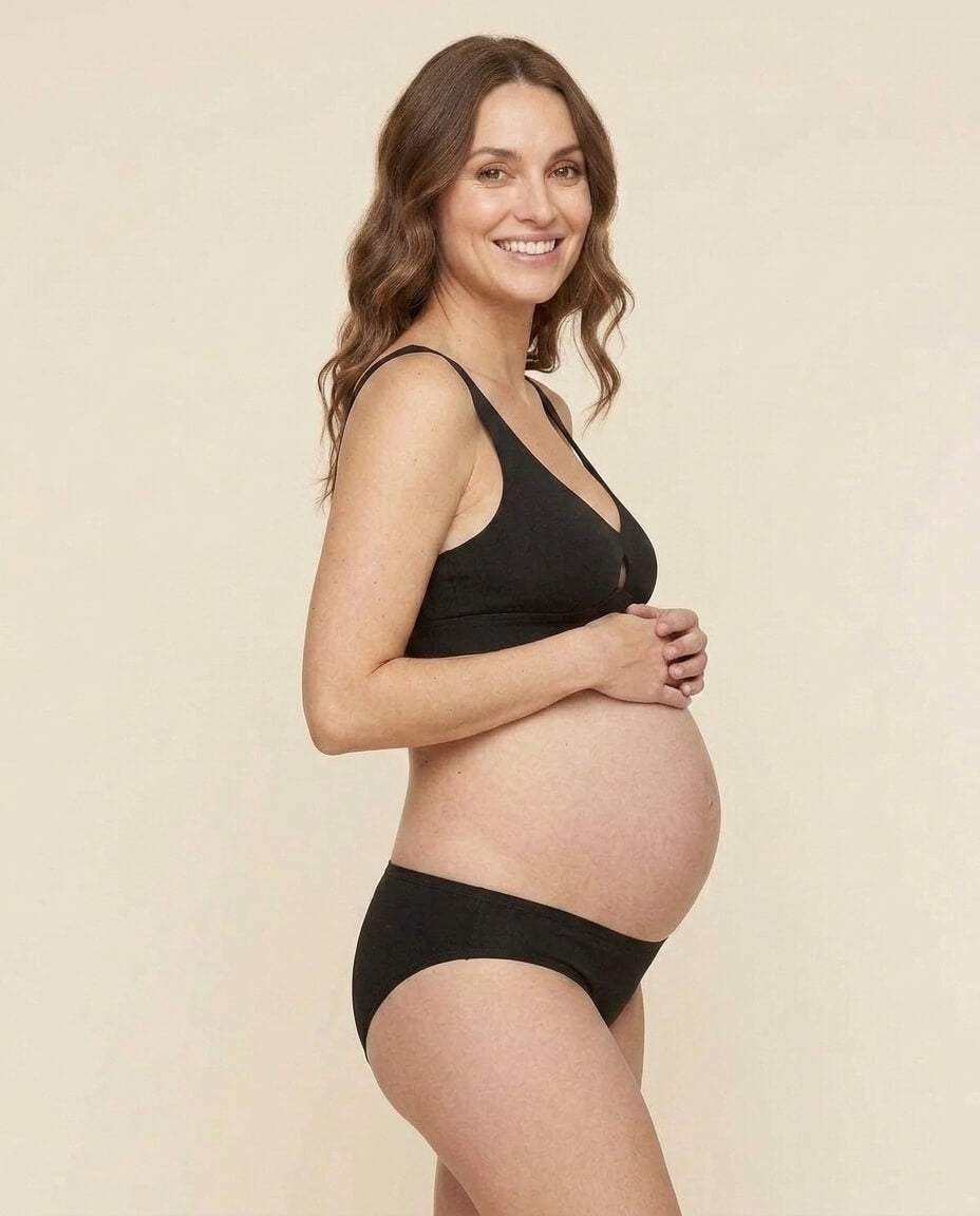 Woman wearing the keyhole soft bra in carbon black and below brief  in carbon black color against a solid light beige background.
