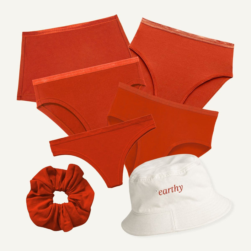 A coordinated apparel set on a light warm-gray background: five pairs of underwear in red-orange shades, a matching red scrunchie, and a white bucket hat with the word 'earthy' in red. No person or real environment is present, so this is Product/Packaging.